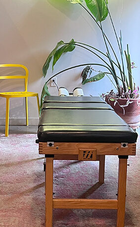Chiropractic bed with yellow chair and a plant in the background.